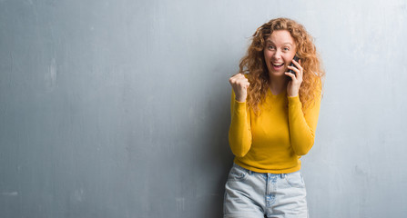 Fototapeta premium Young redhead woman over grey grunge wall talking on the phone screaming proud and celebrating victory and success very excited, cheering emotion