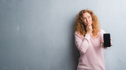 Young redhead woman over grey grunge wall showing smartphone screen serious face thinking about...