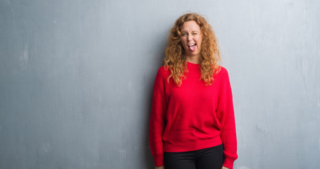 Young redhead woman over grey grunge wall wearing red sweater sticking tongue out happy with funny expression. Emotion concept.