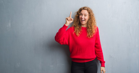 Young redhead woman over grey grunge wall wearing red sweater pointing finger up with successful idea. Exited and happy. Number one.