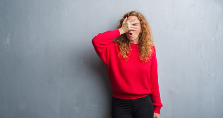 Young redhead woman over grey grunge wall wearing red sweater peeking in shock covering face and...