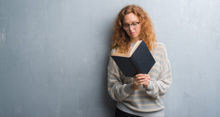 Young redhead woman over grey grunge wall reading a book with a confident expression on smart face thinking serious