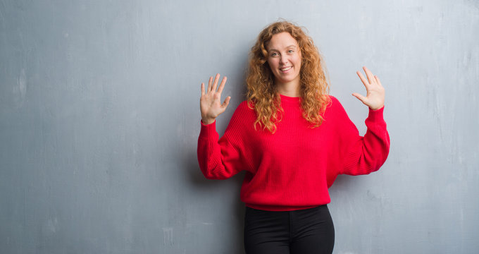 Young redhead woman over grey grunge wall wearing red sweater showing and pointing up with fingers number ten while smiling confident and happy.