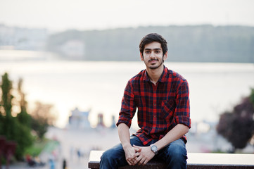 Young indian student man at red checkered shirt and jeans posed at city.