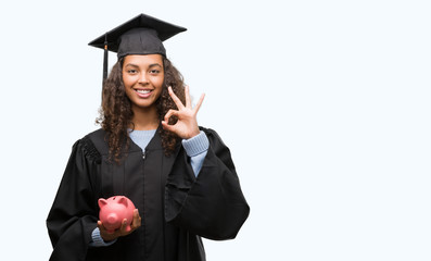 Young hispanic woman wearing graduation uniform holding piggy bank doing ok sign with fingers, excellent symbol