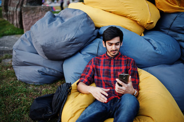 Fototapeta premium Young indian student man at checkered shirt and jeans sitting and relax at outdoor pillows. Spending time with mobile phone.