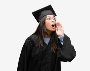 Young hispanic woman wearing graduated cap and uniform shouting and screaming loud to side with hand on mouth. Communication concept.