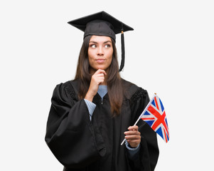 Young hispanic woman wearing graduated uniform holding flag of united kingdom serious face thinking about question, very confused idea