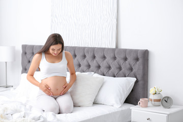 Young beautiful pregnant woman sitting on bed and touching her belly at home