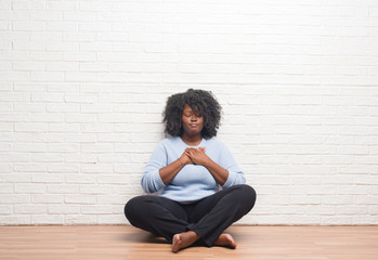 Young african american woman sitting on the floor at home smiling with hands on chest with closed eyes and grateful gesture on face. Health concept.