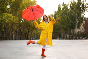 Happy young woman with red umbrella wearing yellow raincoat on street