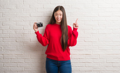 Fototapeta na wymiar Young Chinese woman over brick wall holding vintage camera surprised with an idea or question pointing finger with happy face, number one
