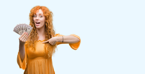 Young redhead woman holding dollars with surprise face pointing finger to himself