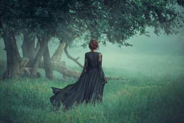 girl with a red hair walking along from dark forest, wearing long black dress with trailer which is...