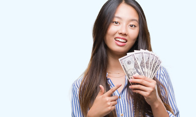 Young asian woman holding dollars over isolated background very happy pointing with hand and finger