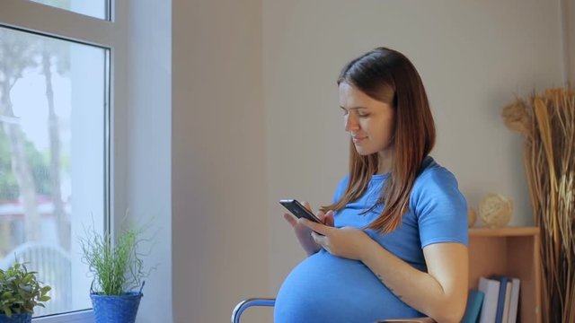 Pregnant woman texting on a mobile on line phone sitting on a sofa in the living room at home.