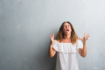 Middle age hispanic woman standing over grey grunge wall crazy and mad shouting and yelling with aggressive expression and arms raised. Frustration concept.