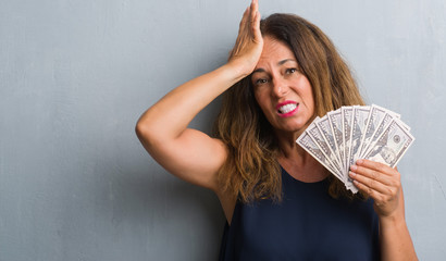 Middle age hispanic woman standing over grey grunge wall holding dollars stressed with hand on head, shocked with shame and surprise face, angry and frustrated. Fear and upset for mistake.