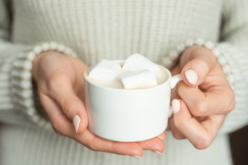 The girl in warm sweaters is holding a white mug with coffee and marshmallow in her hands. A layout for the design of winter gifts, Christmas and New Year.