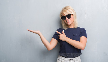 Adult caucasian woman over grunge grey wall wearing sunglasses amazed and smiling to the camera while presenting with hand and pointing with finger.