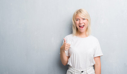 Adult caucasian woman over grunge grey wall doing happy thumbs up gesture with hand. Approving expression looking at the camera with showing success.