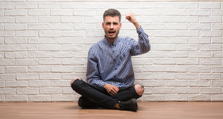 Young adult man sitting over white brick wall angry and mad raising fist frustrated and furious while shouting with anger. Rage and aggressive concept.