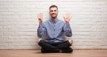 Young adult man sitting over white brick wall showing and pointing up with fingers number nine while smiling confident and happy.