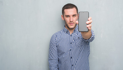Young caucasian man over grey grunge wall showing blank screen of smartphone with a confident expression on smart face thinking serious