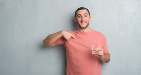 Young caucasian man over grey grunge wall drinking a glass of water with surprise face pointing finger to himself