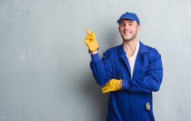 Young caucasian man over grey grunge wall wearing mechanic uniform very happy pointing with hand and finger to the side