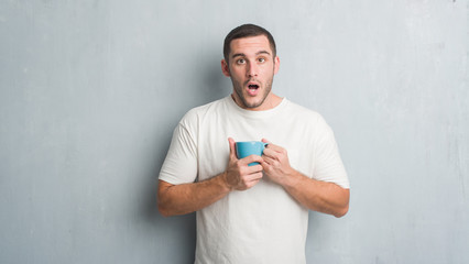 Young caucasian man over grey grunge wall driking a cup of coffee scared in shock with a surprise...