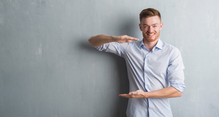 Young redhead business man over grey grunge wall gesturing with hands showing big and large size sign, measure symbol. Smiling looking at the camera. Measuring concept.