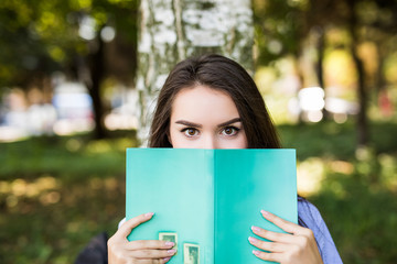 Beautiful girl covering her face with book in summer park backround. Education and people concept