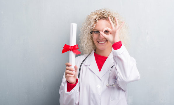 Young blonde doctor woman over grunge grey wall holding degree with happy face smiling doing ok sign with hand on eye looking through fingers