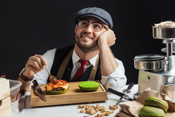 Sweet dreams. Pleasant caucasian man looking up with dreamy expression adding chopped tomatoes to self made burger. His thought are far away from kitchen and food