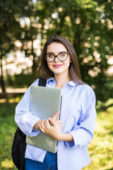 Fototapeta premium Smiling brunette student wearing glasses and shirt walking outdoor with laptop and backpack background of green park