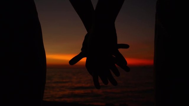 SLOW MOTION, CLOSE UP, SILHOUETTE: Young couple on summer vacation holds hands while watching the picturesque orange sunset. Boyfriend and girlfriend touch and hold hands on a beautiful summer evening