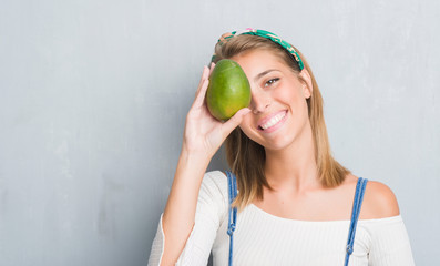 Beautiful young woman over grunge grey wall holding fresh mango with a happy face standing and...