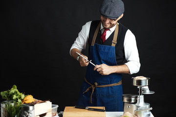 Portrait of chef cook in elegant uniform wearing cap and apron sharpens knife at the restaurant kitchen table over black background. Small business, Service and Catering Concept