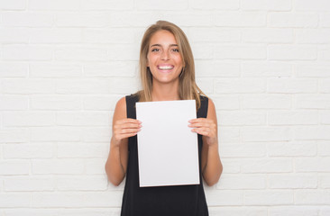 Beautiful young woman over white brick wall holding blank paper sheet with a happy face standing...