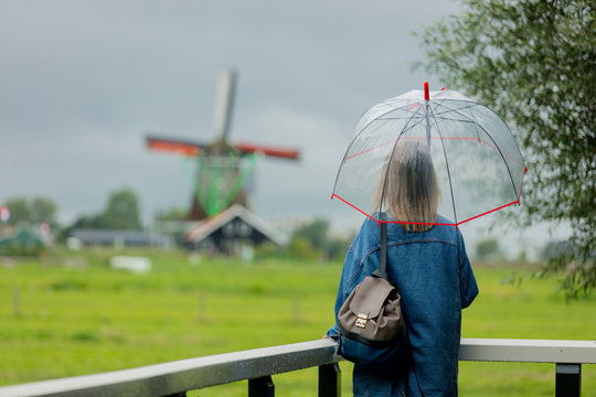 Young girl in jeans jacket with umbrella stay on bridge with dutch mills on background. Netherlands