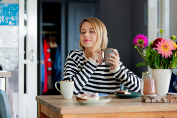 Young woman with cup of coffee or tea and cake sitting in a chair at kitchen in breakfast time. Interior in dutch style.