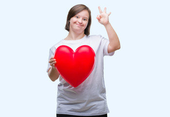 Young adult woman with down syndrome holding red heart over isolated background doing ok sign with fingers, excellent symbol