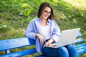 Beautiful young woman in eyesglasses sitting in public park chatting over laptop computer