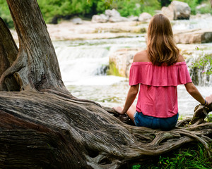 Teen Girl With Long Brown Hair Sitting On A Large Tree Branch With Waterfall In The Background