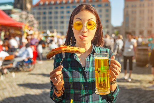 Young happy asian traveler woman eating fried sausage and drinks mug of beer at the fair market square in Germany, beer and food festival concept