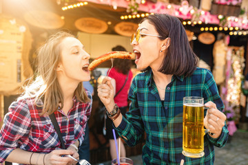 Two girls friends having fun and eating fried sausage and drinks mug of beer at the fair market square in Germany, beer and local food festival concept