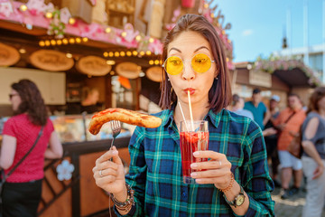 Young happy asian traveler woman eating fried sausage and drinks mug of beer at the fair market...