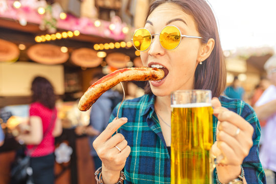Young happy asian traveler woman eating fried sausage and drinks mug of beer at the fair market square in Germany, beer and food festival concept