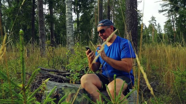 a tourist in sunglasses and a baseball cap in the forest sits on a fallen tree and communicates in the phone in a messenger.